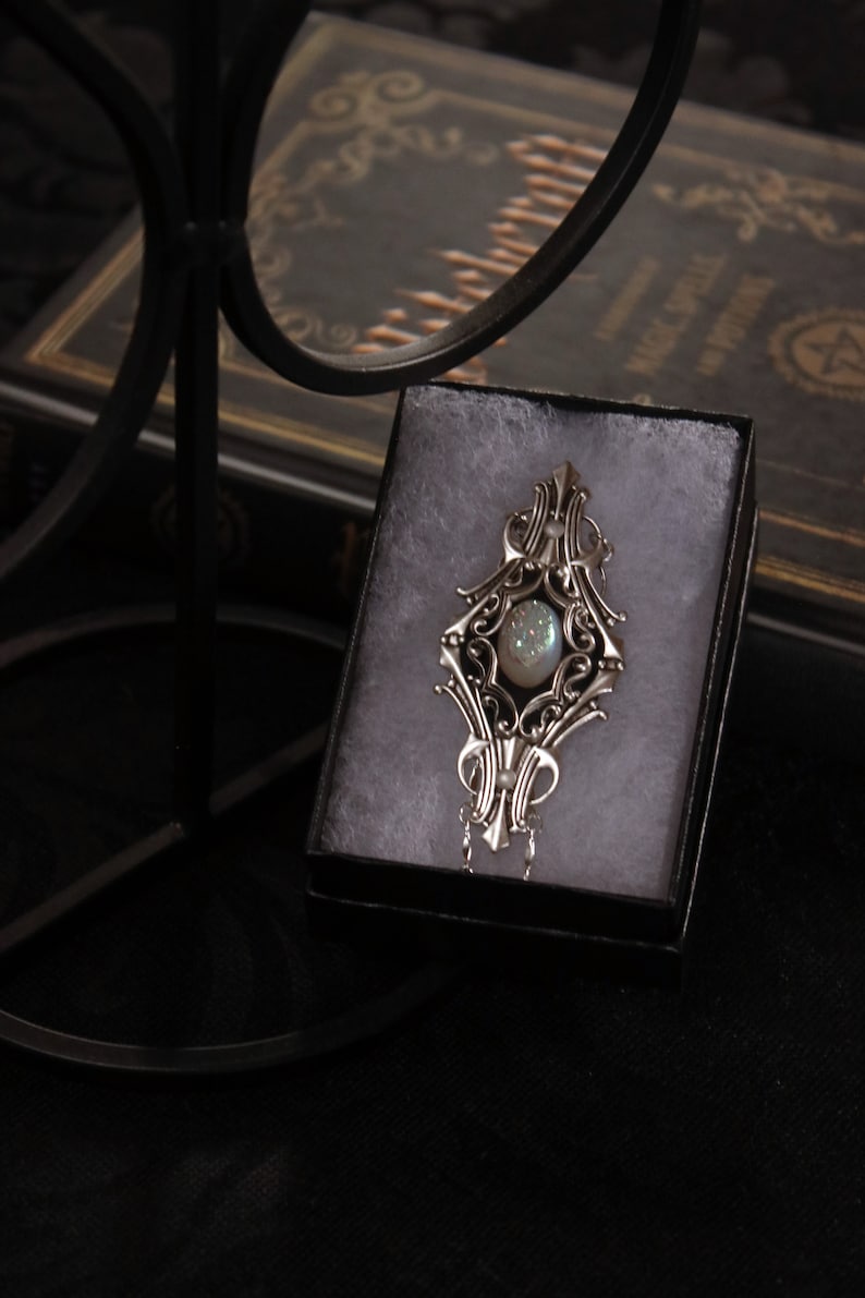 May include: Ornate silver pendant with a central iridescent stone, displayed in a black gift box. The pendant features intricate filigree detailing and a delicate chain. The background includes a dark book, suggesting a mystical or gothic theme.