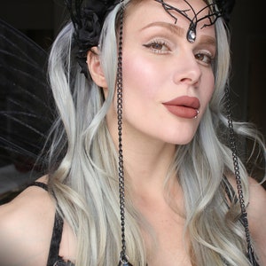 May include: A woman with silver hair wearing a black crown with black roses and feathers. The crown has a chain with a crystal pendant hanging from it. She is wearing a black dress with a lace pattern.