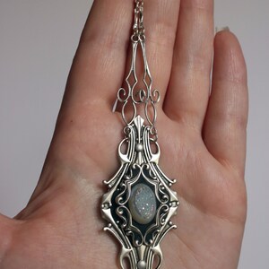 May include: Silver-toned pendant necklace with an ornate, Art Nouveau-inspired design. The pendant features a central oval-shaped stone with a light blue, crystalline appearance. The necklace is held in a hand against a plain background.