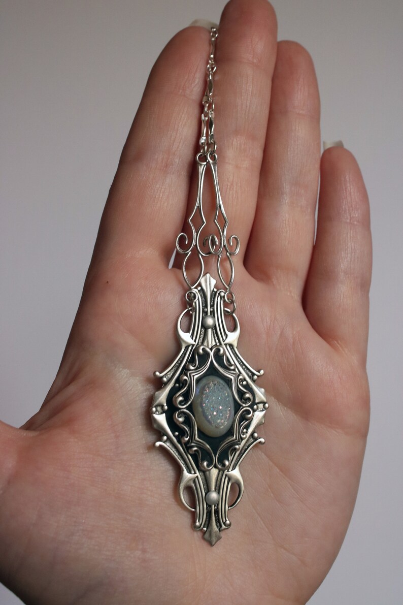 May include: Silver-toned pendant necklace with an ornate, Art Nouveau-inspired design. The pendant features a central oval-shaped stone with a light blue, crystalline appearance. The necklace is held in a hand against a plain background.
