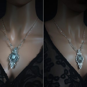 May include: Silver-toned pendant necklace with an ornate, symmetrical design. The pendant features a central, oval-shaped stone and intricate filigree detailing. The necklace is displayed on a person wearing a black lace top.
