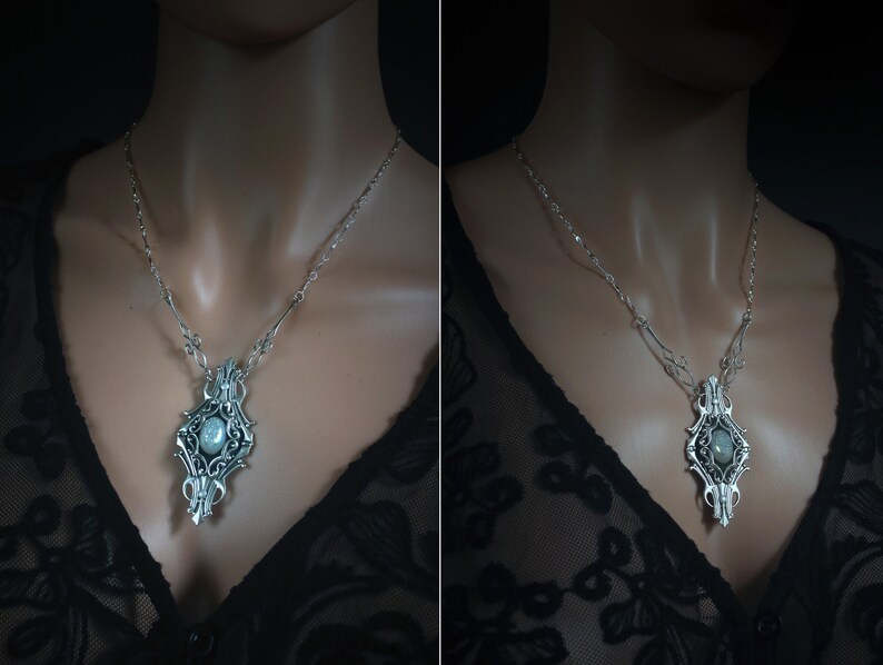 May include: Silver-toned pendant necklace with an ornate, symmetrical design. The pendant features a central, oval-shaped stone and intricate filigree detailing. The necklace is displayed on a person wearing a black lace top.