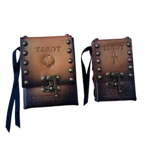May include: Two brown leather tarot card carrying cases with metal accents and clasps. The larger case has the word "TAROT" embossed on the front and a small image of an ankh. The smaller case has the word "TAROT" embossed on the front and a small image of an ankh.