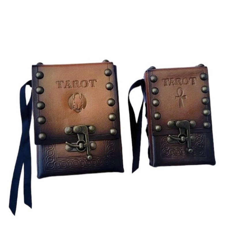 May include: Two brown leather tarot card carrying cases with metal accents and clasps. The larger case has the word "TAROT" embossed on the front and a small image of an ankh. The smaller case has the word "TAROT" embossed on the front and a small image of an ankh.
