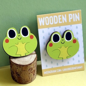 Puede incluir: Un pin de madera verde en forma de rana con ojos negros y mejillas rojas. El pin está sobre un pequeño tronco de madera. El fondo es de color verde claro. El texto "WOODEN PIN" está sobre un fondo amarillo. El texto "INSTAGRAM.COM/JAKUBSOUNFUNNY" está sobre un fondo blanco.