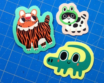 May include: Three colourful animal stickers on a blue cutting mat. The stickers are of a tiger, a raccoon wearing a frog costume, and a green alligator.