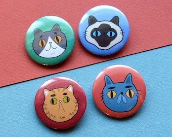 Könnte beinhalten: Vier runde Anstecknadeln mit verschiedenen Katzenköpfen. Die Nadeln sind grün, blau, rot und orange. Jede Katze hat einen anderen Ausdruck und eine andere Farbe.