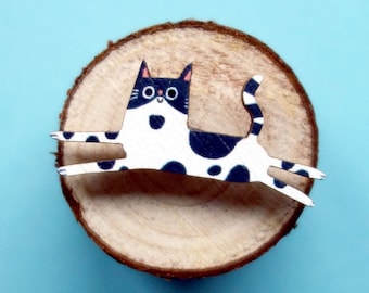 Puede incluir: Un gato blanco y negro con manchas, nariz rosa y ojos negros está recortado en papel y colocado sobre un tronco de madera. El gato está en una pose de salto con las patas extendidas.