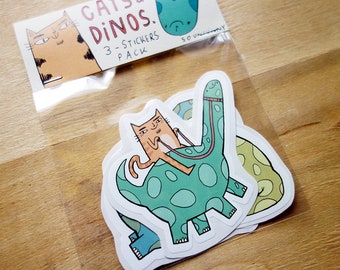 May include: A set of three stickers featuring a cat riding a dinosaur. The stickers are white with a green dinosaur and an orange cat. The text "Cats & Dinos. 3-Sticker Pack" is on the top of the sticker sheet.