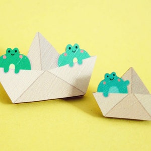 May include: Two paper boats with green frog figures on top. The boats are folded from beige paper and have a simple design. The frogs have smiling faces and are facing forward.
