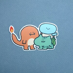 May include: A sticker featuring three cartoon characters: a red and orange fire-breathing dragon, a blue and white cloud-like creature, and a green frog-like creature.