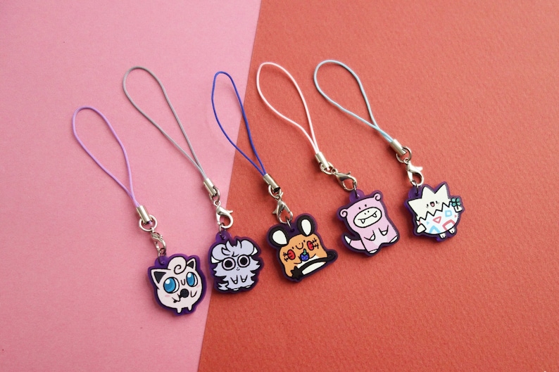 Op de afbeelding: Vijf kleurrijke cartoon karakter bedels met bijpassende gekleurde koorden en zilveren sluitingen. De bedels zijn paars, wit, oranje en roze, met verschillende cartoon karakter ontwerpen. De achtergrond is roze en rood.