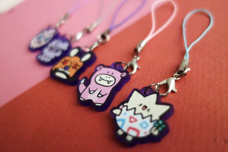 Op de afbeelding: Een verzameling kleurrijke cartoonkarakterbedels met zilveren sluitingen en bijpassende lanyards. De bedels hebben verschillende ontwerpen, waaronder een paars konijn, een roze wezen en een wit en blauw eivormig karakter. De achtergrond is een mix van roze en rood.