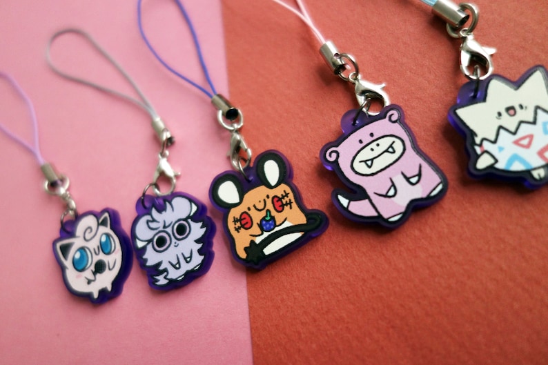 Op de afbeelding: Een verzameling kleurrijke cartoon-charms met paarse randen en bevestigde koorden. De charms hebben verschillende ontwerpen, waaronder een roze wezen, een wit en paars wezen en een bruin en oranje wezen. De achtergrond is een mix van roze en rood.