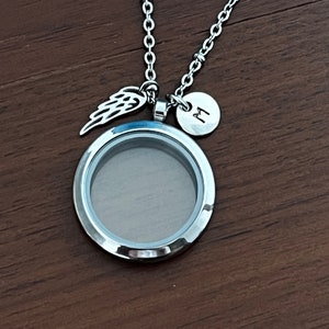 May include: A silver chain necklace with a round locket, a small angel wing charm, and a round charm with the letter "M" engraved on it.