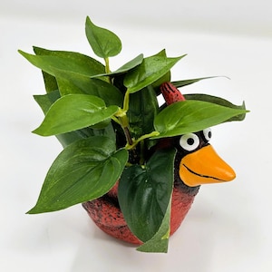 Könnte beinhalten: Ein roter, vogelförmiger Pflanztopf mit schwarzem und orangefarbenem Schnabel, der grüne Blätterpflanzen enthält. Der Pflanztopf hat eine strukturierte Oberfläche und ist mit leuchtend grünem Laub gefüllt, was eine dekorative und skurrile Darstellung ergibt.