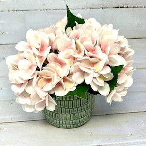 May include: A cluster of pale pink hydrangea flowers with green leaves arranged in a round, textured sage green ceramic pot. The petals feature soft white and light pink gradients, while the pot displays a repeating vertical grid pattern.