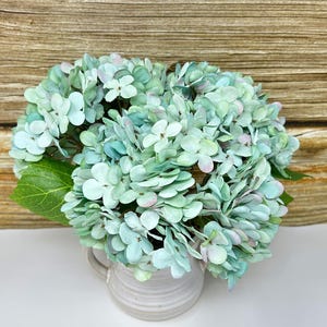 May include: A bouquet of artificial hydrangeas in shades of blue, green, and pink, arranged in a small, off-white ceramic pitcher. The flowers have multiple layers of petals, and a single green leaf is visible. The background is a wooden surface.