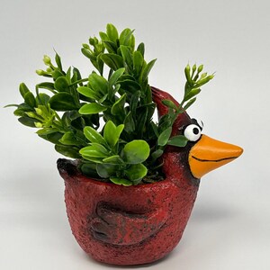 Könnte beinhalten: Ein roter, keramik Vogel-Blumentopf mit orangefarbenem Schnabel und weißen Augen, gefüllt mit grünem Kunstlaub. Der Topf hat eine strukturierte Oberfläche und ist als Kardinalvogel gestaltet. Ein dekorativer Artikel für die Inneneinrichtung.