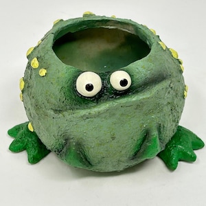 Puede incluir: Una maceta con forma de rana verde caprichosa, con grandes detalles de ojos blancos y manchas amarillas. La maceta de cerámica tiene una superficie texturizada y pequeños pies verdes. Ideal para plantas pequeñas.