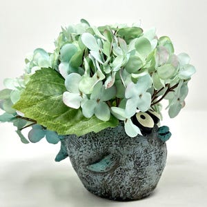 May include: A decorative floral arrangement featuring light green and blue artificial hydrangeas and leaves. The flowers are arranged in a textured, blue-gray ceramic pot with a whimsical bird design.