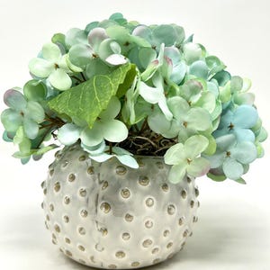 May include: A decorative floral arrangement featuring a cluster of green and blue hydrangea blooms. The flowers are arranged in a round, textured, off-white ceramic vase with small, gold-colored dot accents. A green leaf is visible.