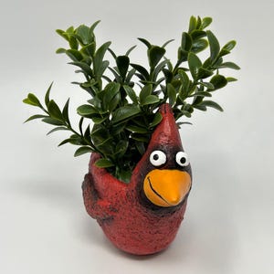 Puede incluir: Una maceta roja con forma de cardenal, con una expresión alegre, pico naranja brillante y ojos blancos. La maceta está llena de follaje artificial verde vibrante, creando un acento decorativo lúdico.