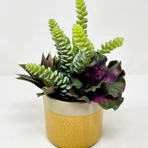 Pode incluir: Um pequeno arranjo decorativo de suculentas num vaso de cerâmica texturizada amarelo e bege. O arranjo apresenta plantas artificiais verdes e roxas, criando um contraste vibrante. O vaso tem cerca de 10 cm de altura.
