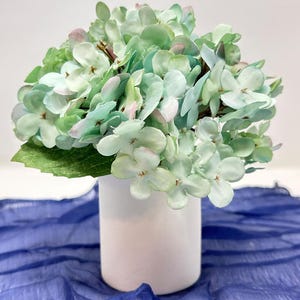 May include: A bouquet of artificial hydrangea flowers in shades of mint green and pale pink, arranged in a simple white cylindrical vase. The flower petals have a soft, rounded appearance, and a single green leaf is visible at the base.