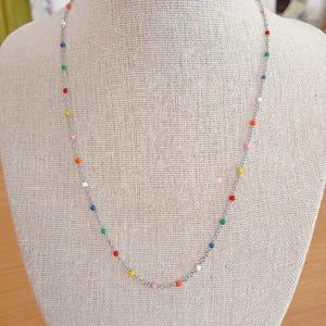 Può includere: Una delicata collana a catena color argento con piccole perline smaltate multicolori distanziate uniformemente nei toni del rosso, blu, verde, giallo, arancione, bianco e rosa, creando un accessorio vivace e semplice.