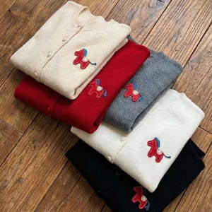 May include: A stack of button-down cardigans in various colors: cream, red, gray, white, and black. Each sweater features an embroidered red horse design with blue accents. The sweaters are neatly folded on a wooden surface.