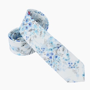 Peut inclure: Une cravate blanche avec un motif floral bleu. La cravate a un effet aquarelle et présente de petites fleurs bleues sur des branches blanches.
