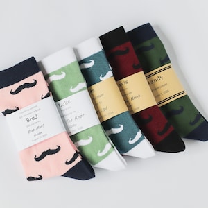 Peut inclure: Cinq paires de chaussettes avec un motif de moustache et une étiquette personnalisée. Les étiquettes sont brunes avec du texte blanc et comprennent les noms Brad, Luke, John, Davis et Randy. Les chaussettes sont de différentes couleurs, notamment rose, vert, bleu, rouge et noir.