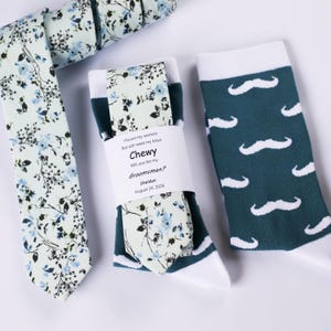 May include: A light green floral tie and socks set with a matching design. One pair of socks has a mustache pattern, and the other has a floral pattern. The socks are packaged with a label that reads "Chewy, Will you be my Groomsman?"