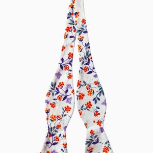 May include: A white bow tie with a floral pattern featuring orange and purple flowers on a white background.