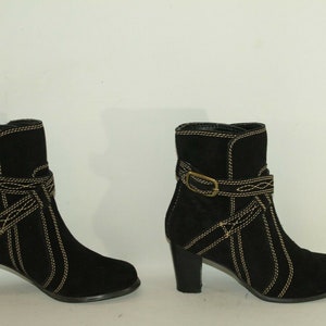 Pode incluir: Um par de botas de tornozelo de camur&ccedil;a preta com costuras douradas e um detalhe de fivela. As botas t&ecirc;m um salto bloco.