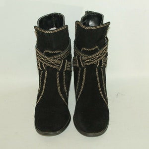 Pode incluir: Um par de botas de tornozelo de camur&ccedil;a preta com detalhes de costura dourada. As botas t&ecirc;m um salto baixo e uma ponta pontiaguda.