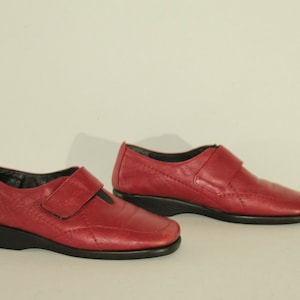 Puede incluir: Un par de zapatos de cuero rojo con una correa y un cierre de hebilla. Los zapatos tienen una suela negra y una punta ligeramente redondeada.