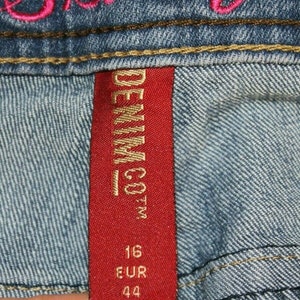 Puede incluir: Etiqueta de mezclilla roja con letras doradas que dicen "DENIM CO.", con los n&uacute;meros "16 EUR 44" impresos debajo.