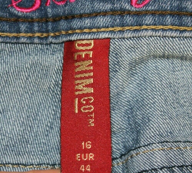 Puede incluir: Etiqueta de mezclilla roja con letras doradas que dicen "DENIM CO.", con los n&uacute;meros "16 EUR 44" impresos debajo.