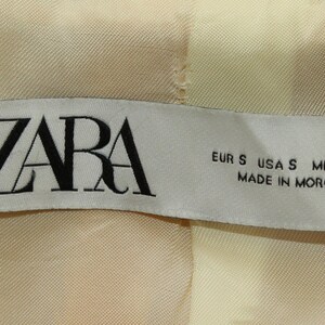 Puede incluir: Etiqueta blanca con texto negro que dice "ZARA" y "EUR S USA S MEX 26 MADE IN MOROCCO".