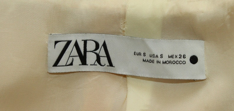 Puede incluir: Etiqueta blanca con texto negro que dice "ZARA" y "EUR S USA S MEX 26 MADE IN MOROCCO".