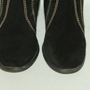 Pode incluir: Um par de botas de tornozelo de camur&ccedil;a preta com costuras douradas. As botas t&ecirc;m uma ponta arredondada e um salto baixo.