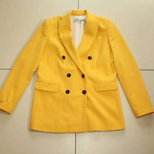 Puede incluir: Una chaqueta amarilla de doble botonadura con botones negros. La chaqueta tiene un corte cl&aacute;sico y est&aacute; hecha de una tela lisa.
