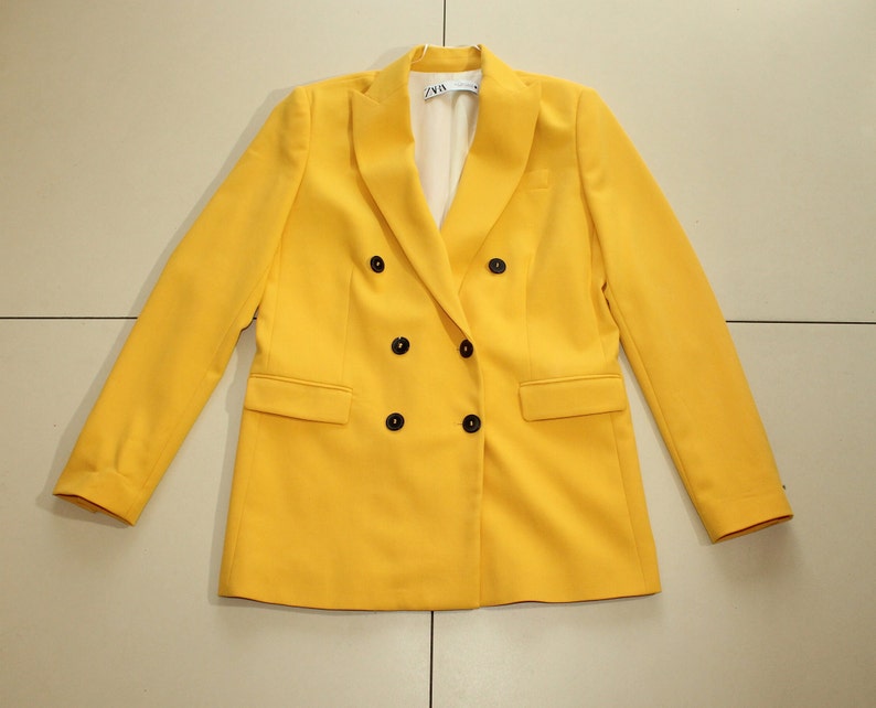 Puede incluir: Una chaqueta amarilla de doble botonadura con botones negros. La chaqueta tiene un corte cl&aacute;sico y est&aacute; hecha de una tela lisa.