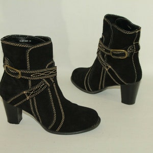 Pode incluir: Botas de tornozelo de camur&ccedil;a preta com fivela dourada e detalhes de costura. As botas t&ecirc;m um salto bloco.