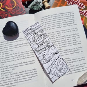 Op de afbeelding: Zwart-witte bladwijzer met een tekening van boeken, geplaatst in een open boek. Een zwarte hartvormige steen is zichtbaar. Het boek heeft tekst in het Duits.