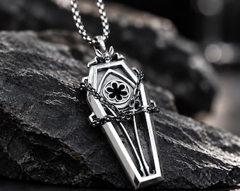 May include: A silver-toned metal pendant shaped like a coffin, featuring a central black flower emblem and a small bat figure at the top. The pendant is wrapped in a thin black chain and hangs from a silver-toned box chain necklace.