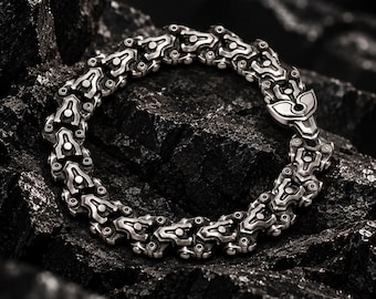 May include: A silver-toned metal chain bracelet featuring a series of repeating, intricate mechanical-style links with recessed dark detailing and a secure clasp mechanism, arranged in a circular shape on a dark, textured surface.