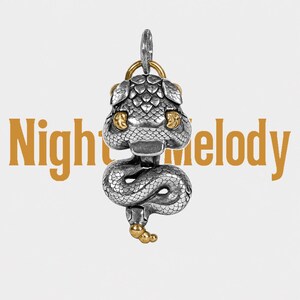 May include: A silver and gold-toned metal pendant shaped like a coiled snake with detailed scales, featuring a prominent head and a segmented tail. The text Night Melody is displayed in large, light brown letters behind the snake-shaped pendant.